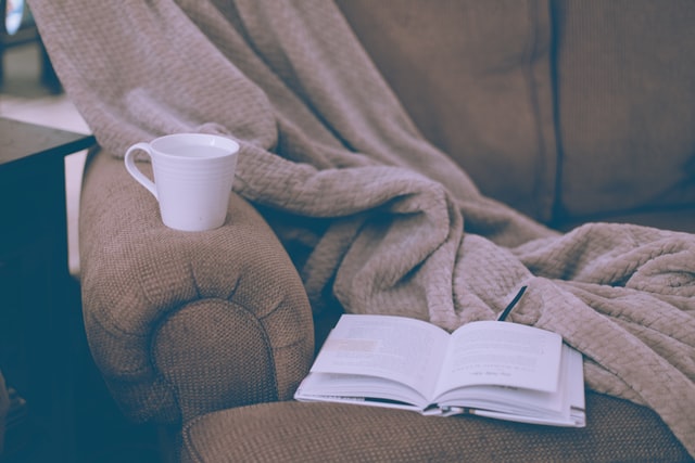 An open book, a white ceramic mug, and a blanket rest upon a couch.